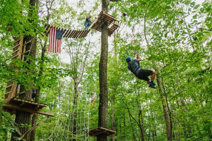 Person zip-lining through a forest adventure park with an American flag hanging nearby.