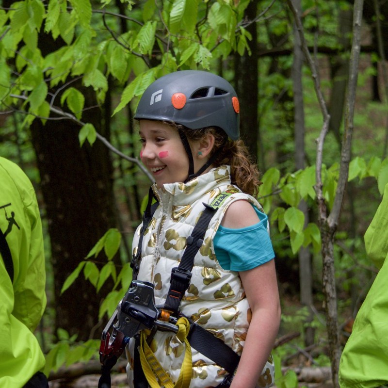 Girl in helmet and vest smiling in a forest with two people in green jackets.