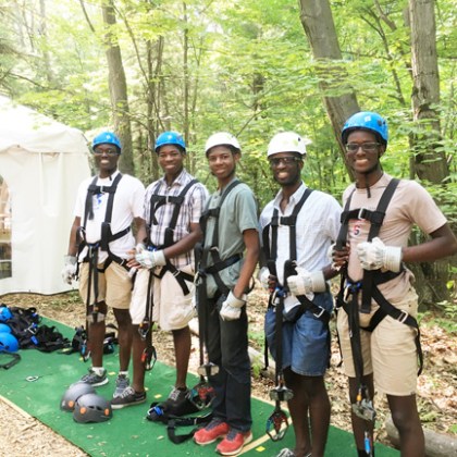 Five people in harnesses, helmets, and gloves stand in a forest near a tent.