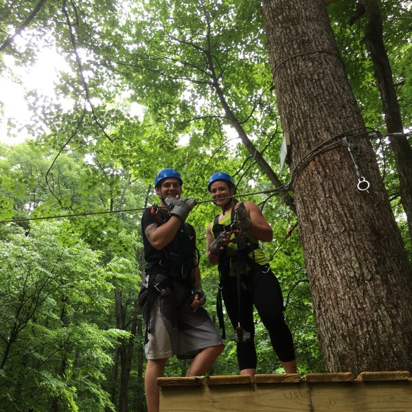 Two people in harnesses stand on a platform in a forest, preparing for zip-lining.