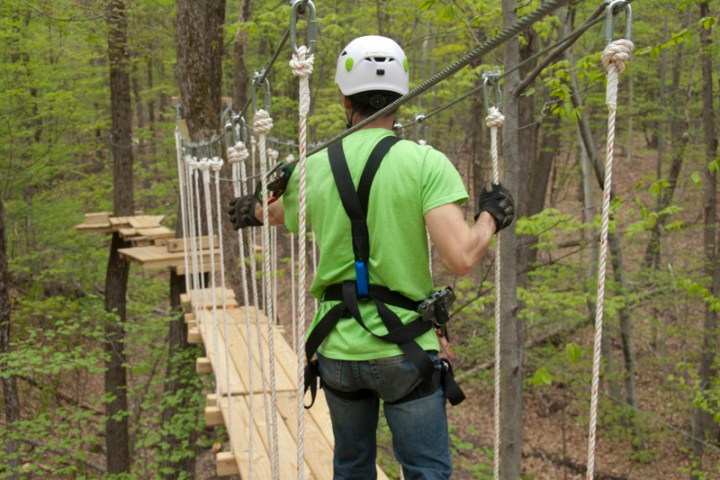 Person in safety gear crossing a rope bridge in a forest.