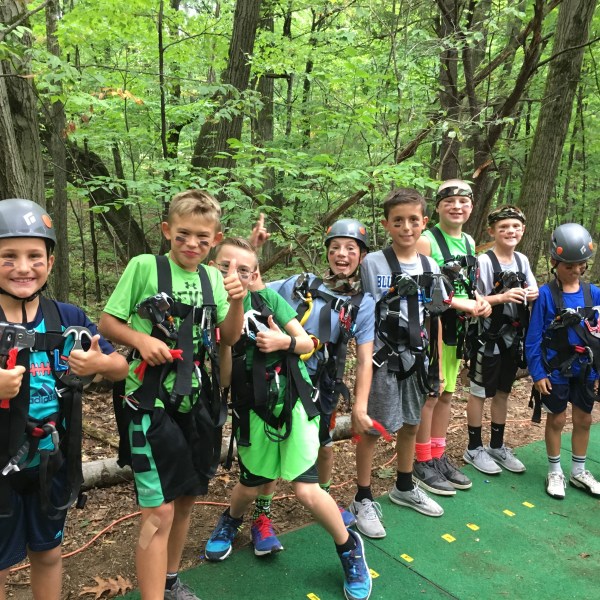 Kids in helmets and gear standing in a forest, smiling and giving thumbs up.