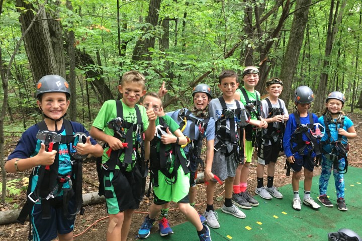 Kids in helmets and gear standing in a forest, smiling and giving thumbs up.