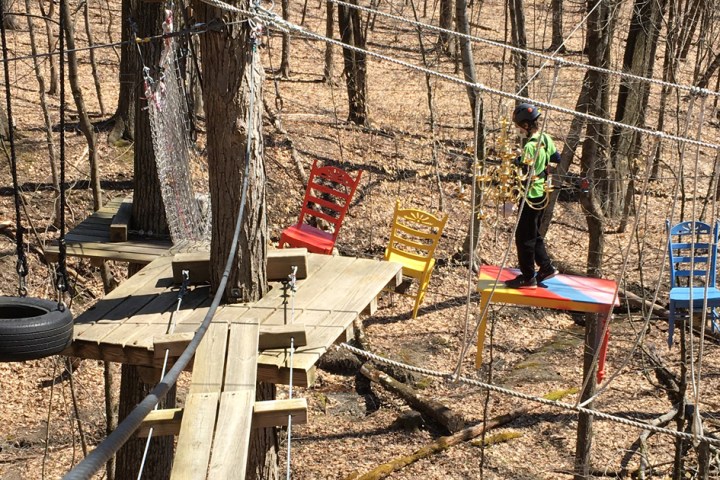 Person on elevated platform in a forest, surrounded by chairs and ropes.