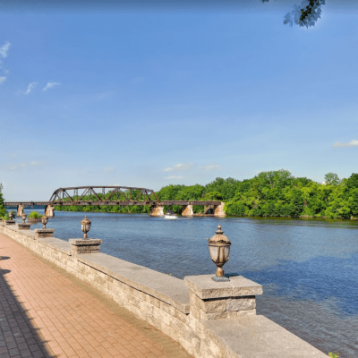 River view with stone walkway, lampposts, and metal bridge under a blue sky.
