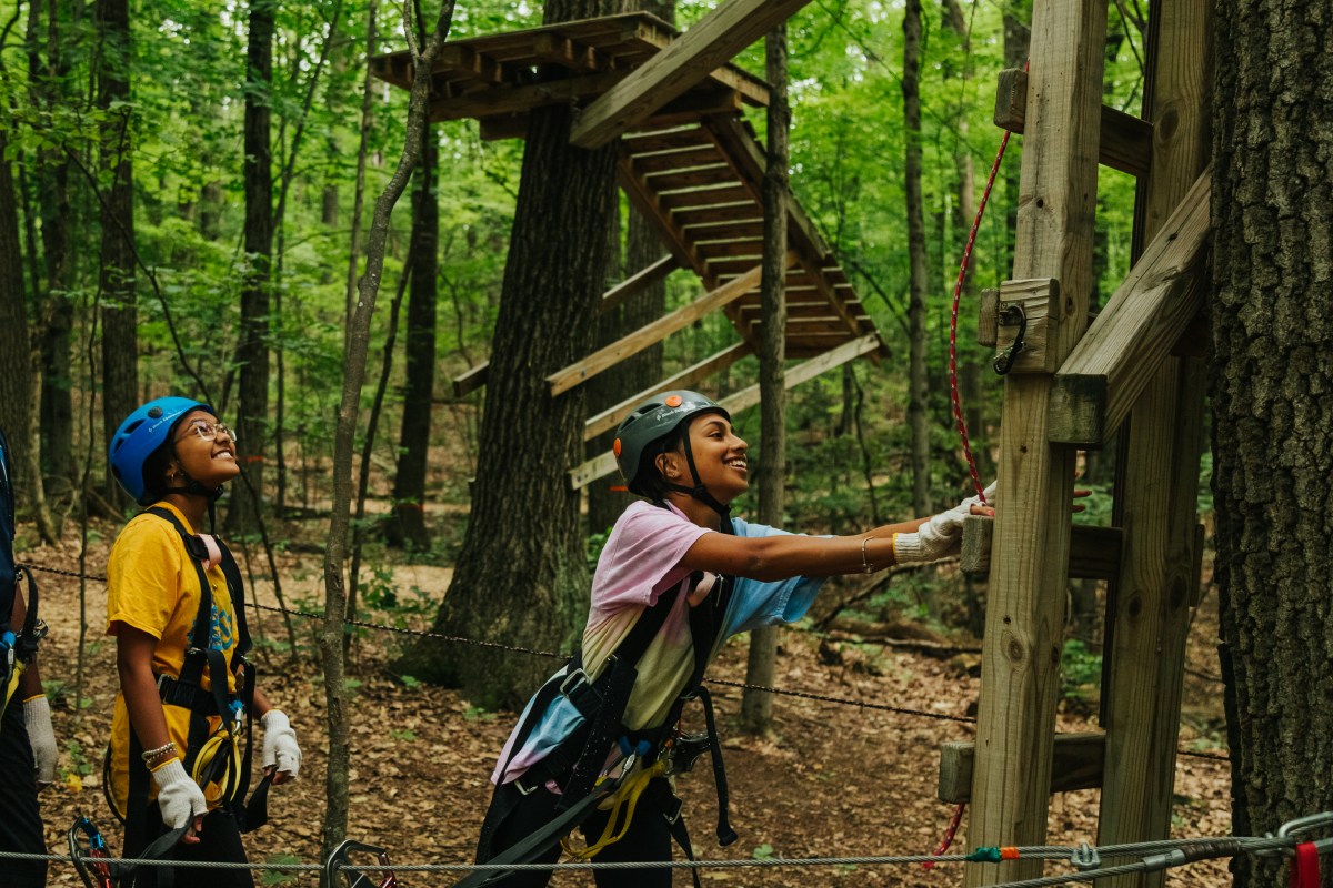 Two kids in helmets preparing for a tree climbing activity in a forest.