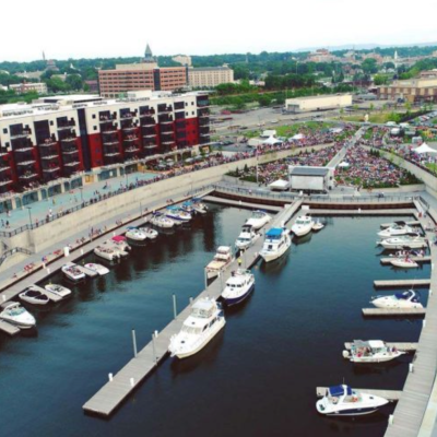 Aerial view of a marina with boats docked and a large crowd gathered in a nearby open space.