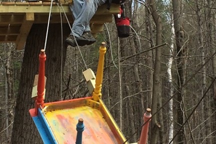 Man sitting on tree platform with colorful upside-down table lifted by ropes.