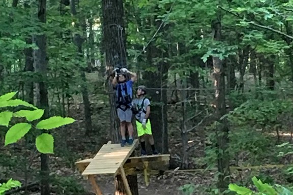 Two people stand on a wooden platform in a forest, preparing for a zipline.