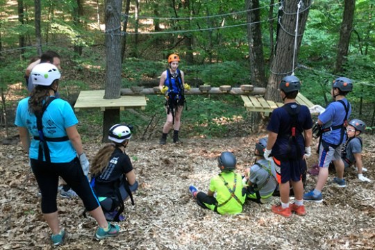 Group of kids in helmets listen to a guide at a ropes course in the woods.