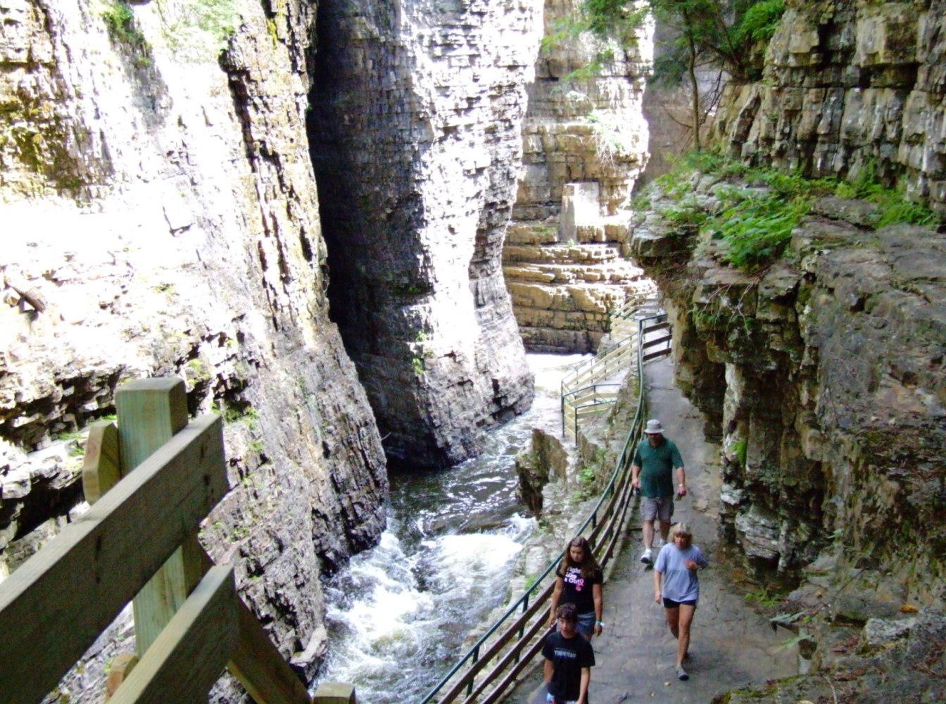 People walking on a narrow path through a rocky gorge with a flowing river.